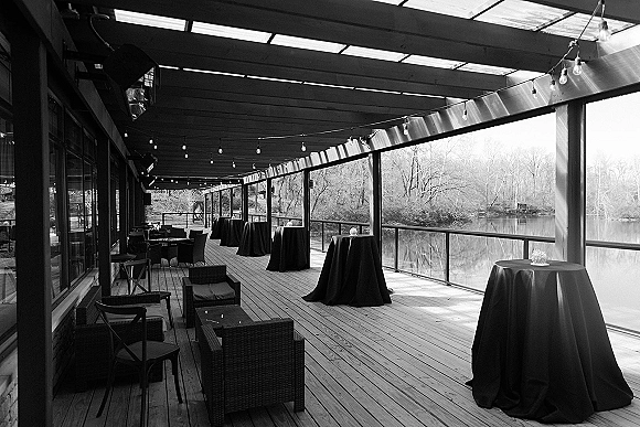 Cocktail hour setup with outdoor cocktail hour high top tables in black linens, small florals, and string lights on a riverfront deck