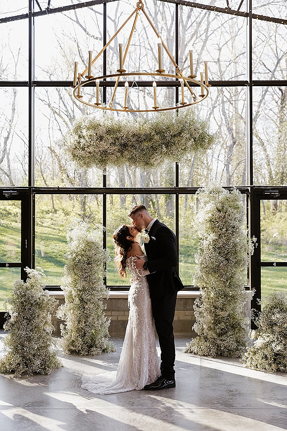 Wedding kiss portrait of bride and groom kissing under a gold chandelier with taper candles and baby’s breath pillars in a glass-walled venue
