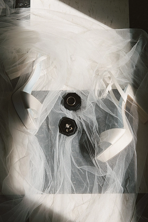 Wedding rings beside an engagement ring flat lay in a black velvet octagon ring box, styled with tulle veil and white bridal heels on fabric