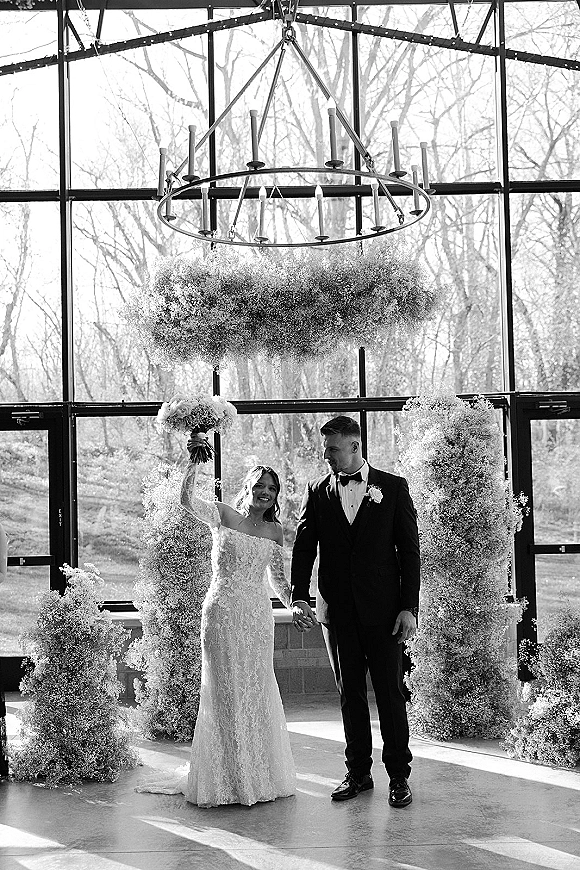 Wedding couple portrait of newlyweds holding hands as the bride lifts a white bouquet beside a glass wall with hanging flowers overhead
