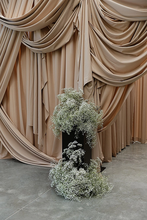 Wedding ceremony backdrop with draped fabric and beige curtains, featuring baby's breath flowers on a black pedestal in an indoor venue