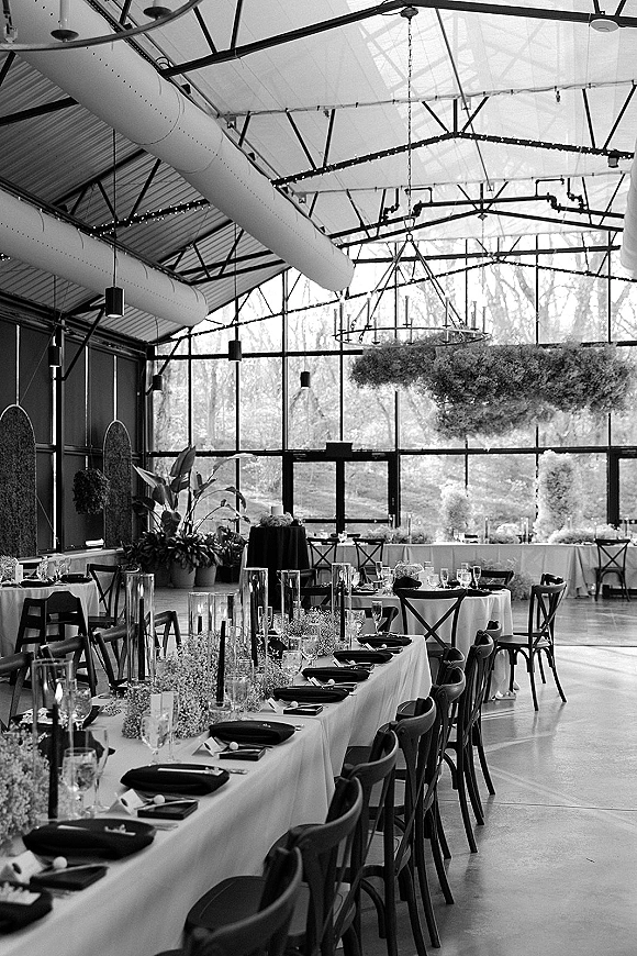 Reception tablescape with long banquet table decor, white linens and black napkins, taper candles and baby's breath beneath greenery chandelier in greenhouse