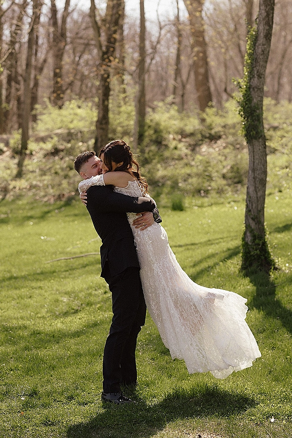 Wedding couple portrait of groom lifting bride in a lace wedding dress with long train, hugging on a grassy lawn among woodland trees