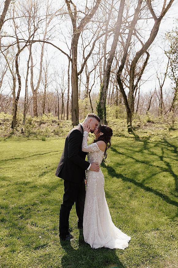 Wedding kiss portrait of bride and groom kissing, her hand on his face, in a black tux and lace-sleeve gown with veil on a woodland lawn