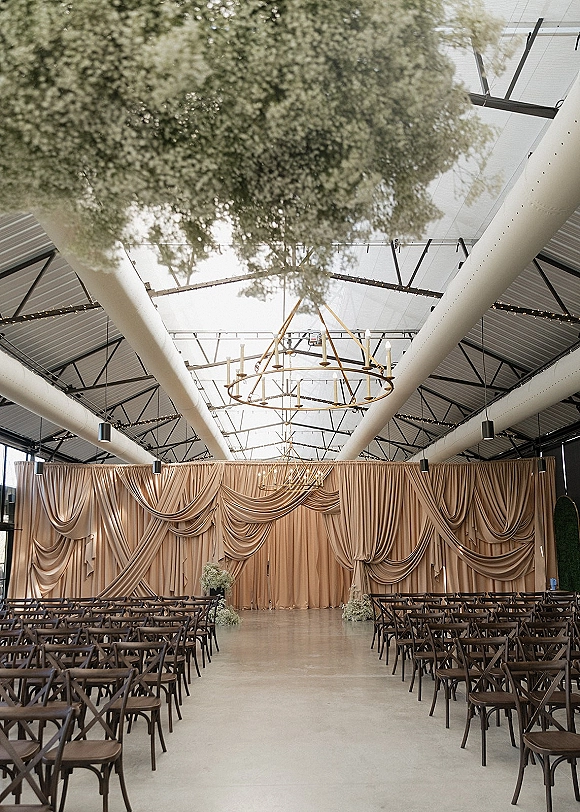 Ceremony setup for indoor wedding ceremony with crossback chairs, draped fabric backdrop, and a chandelier under skylit industrial beams