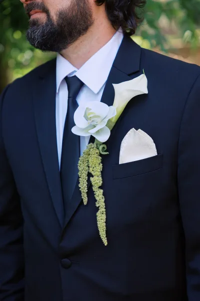 Groom boutonniere with a white calla lily boutonniere and trailing greenery pinned to a black suit, blurred foliage behind