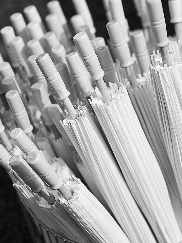 Wedding umbrellas in a basket with white canopies and wooden handles lined up against a dark wall, ready for guests on rainy days