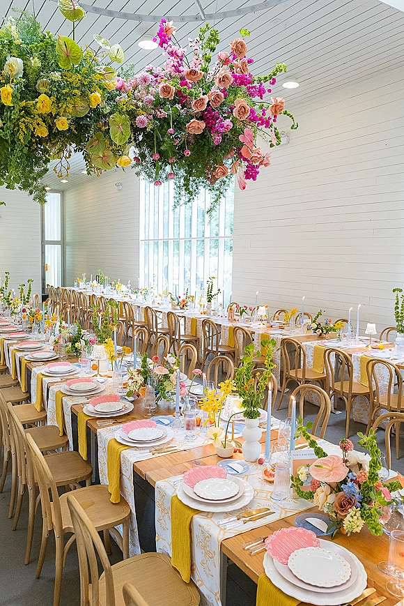 Reception tablescape with hanging floral installation, pink and yellow flowers, taper candles and patterned linens on farmhouse tables by large windows