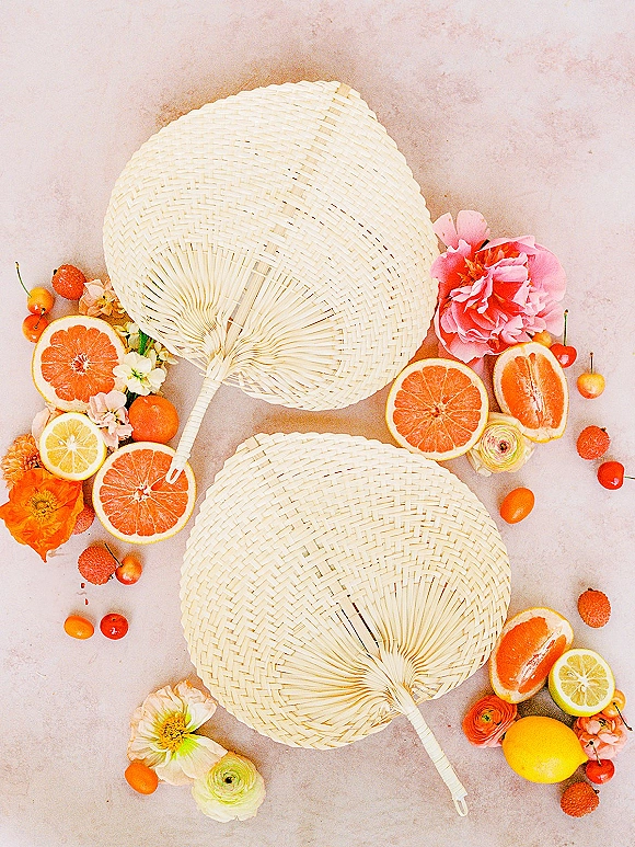 Wedding flat lay of citrus slices, cherries, and ranunculus beside woven hand fans on a neutral stone surface for bright summer details