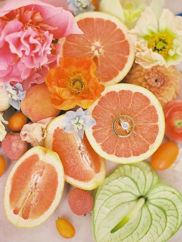 Engagement ring flatlay with grapefruit ring photo styling, featuring citrus slices, kumquats, peach, apple, and flowers on a light tabletop