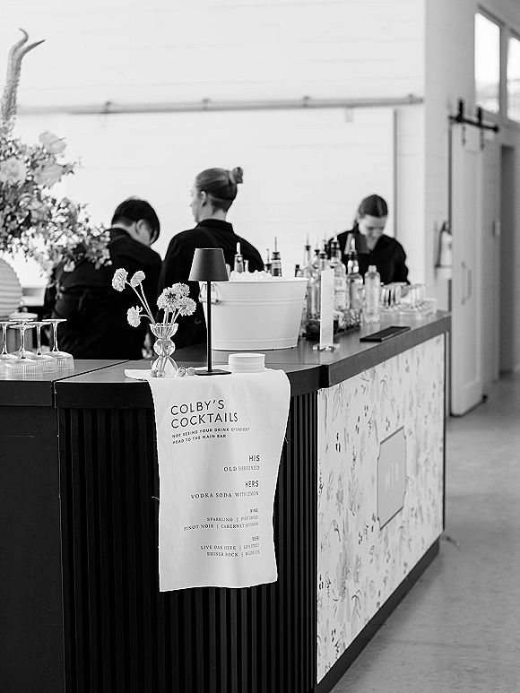 Wedding bar setup with a cocktail menu sign, bottles, glassware, and a small lamp on the counter as staff work behind the bar indoors
