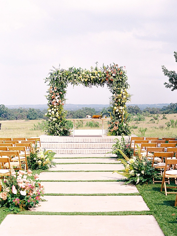 Ceremony setup for an outdoor wedding ceremony with a floral wedding arch, aisle flowers, and wooden chairs in an open field with hills