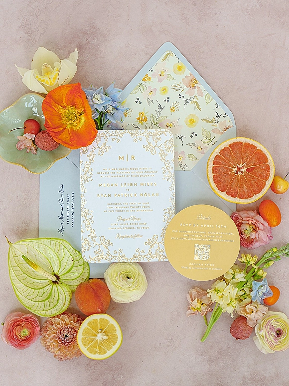 Wedding invitation suite flat lay with gold foil lettering, floral liner, and fruit slices with flowers on a pink stone surface