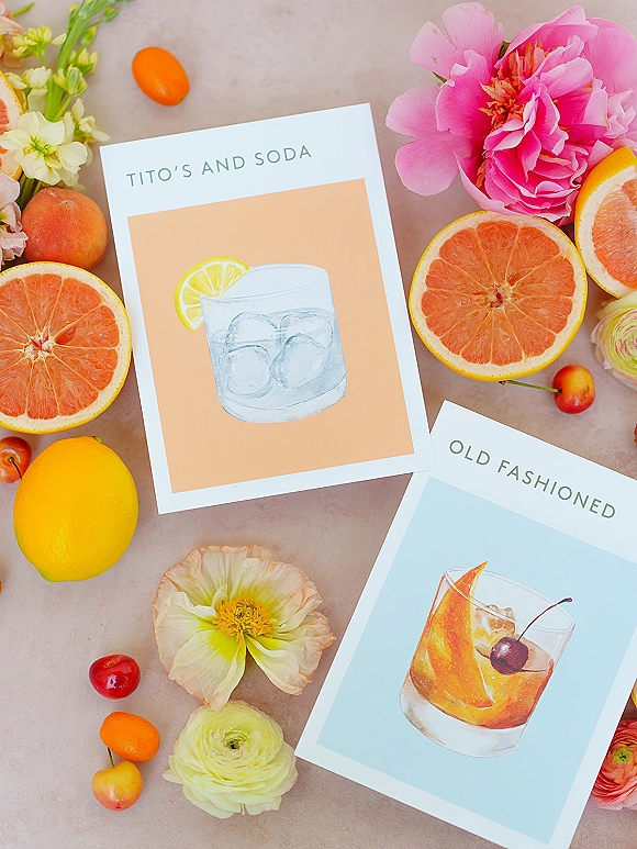 Signature drink sign with wedding bar signage, illustrated cocktail cards beside grapefruit, kumquats, cherries, and peony blooms on tabletop
