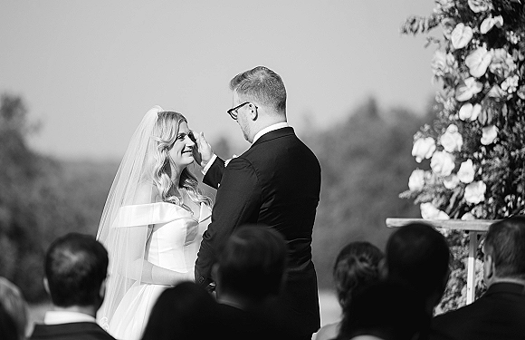 Wedding vows at an outdoor wedding ceremony as the groom wipes the bride’s tear while they hold hands by a floral podium under open sky