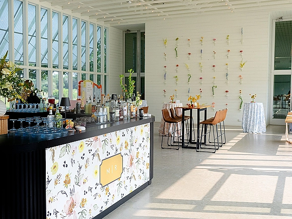 Wedding bar setup with liquor bottles, cocktail glasses, and citrus garnish station on a black counter, lit by string lights and flower wall backdrop
