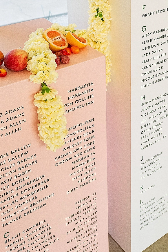 Wedding seating chart escort card display on blush acrylic blocks with guest list cards, yellow floral garland, and citrus fruit on a tabletop