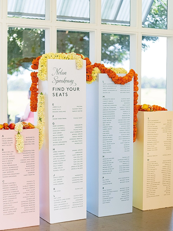 Wedding seating chart and find your seat sign on pastel boards atop pedestals, with marigold garland and citrus accents by sunlit windows