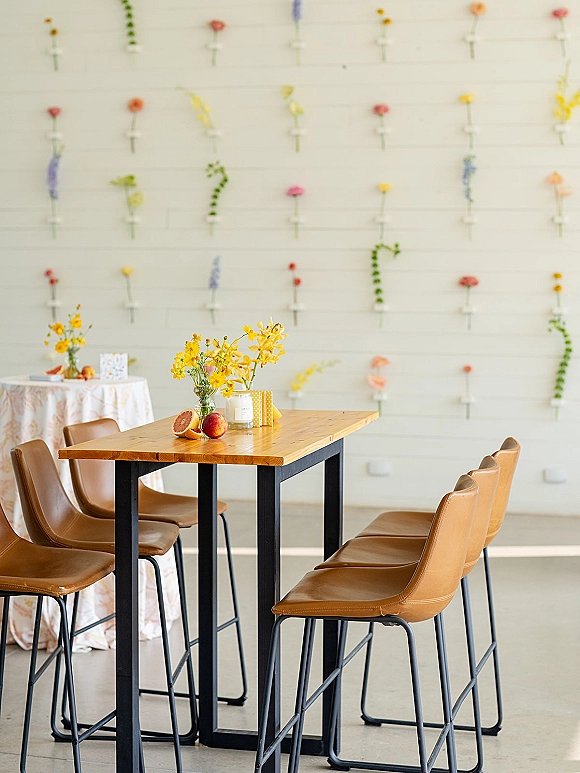 Cocktail table decor with bud vases of yellow flowers, citrus, and a candle on patterned linen, beside bar stools against a hanging flower wall