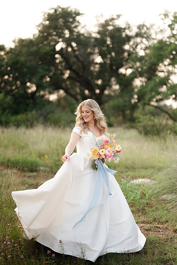 Bridal portrait of a bride holding bouquet in a white off-the-shoulder gown, looking down in a wildflower meadow with trees and sky