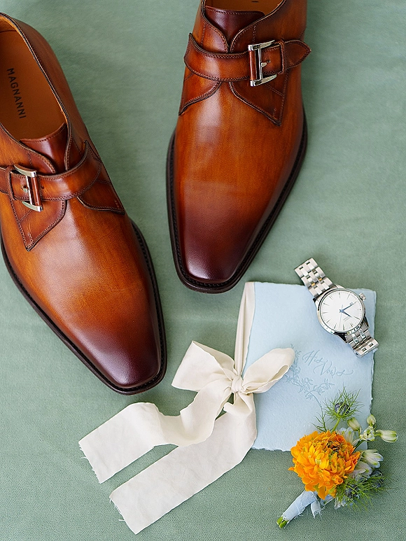 Groom details flat lay with brown leather shoes, silver wristwatch, ribbon-tied vow book and orange boutonniere on green fabric backdrop