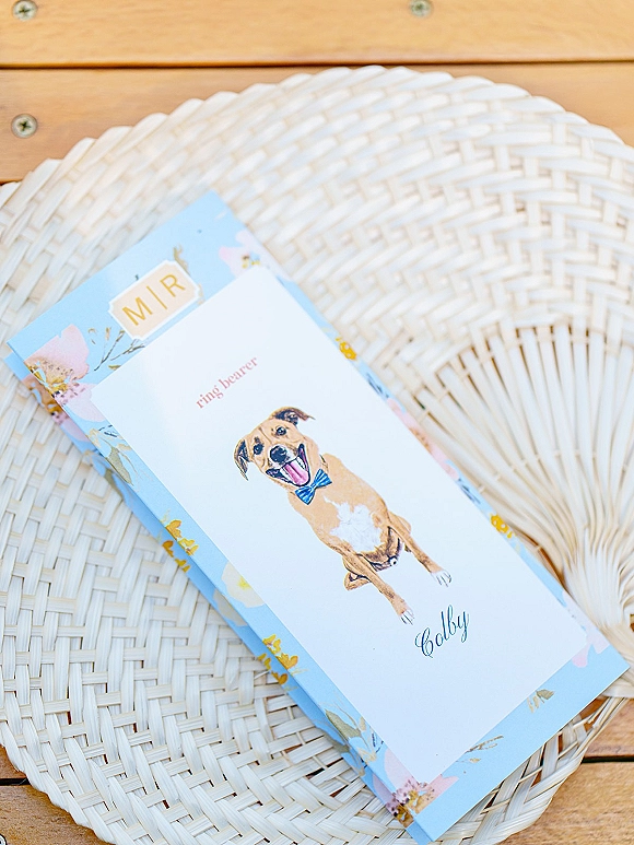 Wedding program with pet wedding program details, featuring an illustrated dog in a bow tie with floral border on a woven fan and wooden table