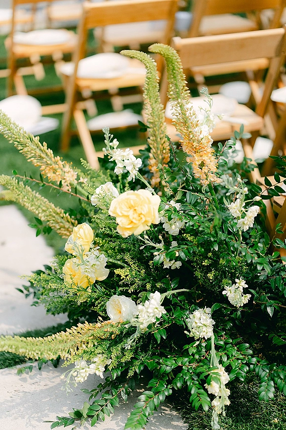 Ceremony aisle flowers with white blooms and yellow roses on a stone walkway, lined by wooden folding chairs with white cushions on grass lawn