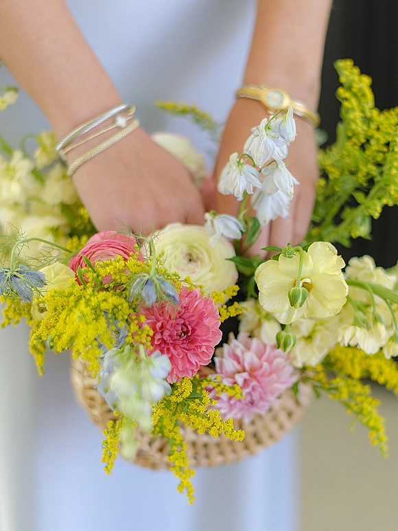 Wedding bouquet in a wicker basket held in front of a white dress and dark suit, filled with mixed pastel flowers and greenery