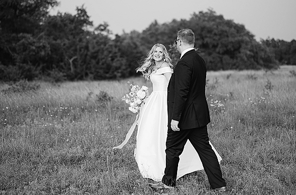 Couple portrait in a black and white wedding photo, bride in off-the-shoulder dress holding ribboned bouquet, walking in wildflower field
