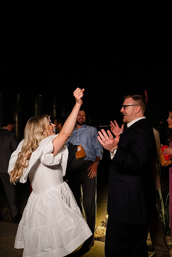 Wedding reception dancing as bride in a short bridal dress and groom in a black suit laugh under the night sky with guests on an outdoor walkway
