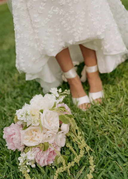 Bridal bouquet with pink peonies and white roses beside the bride’s gown hem and ankle-strap heels on a grass lawn