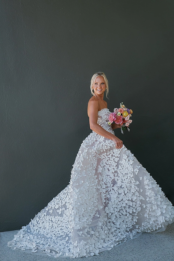 Bridal portrait of a bride in a strapless wedding dress with 3D floral appliqué, holding a colorful bouquet against a dark wall backdrop