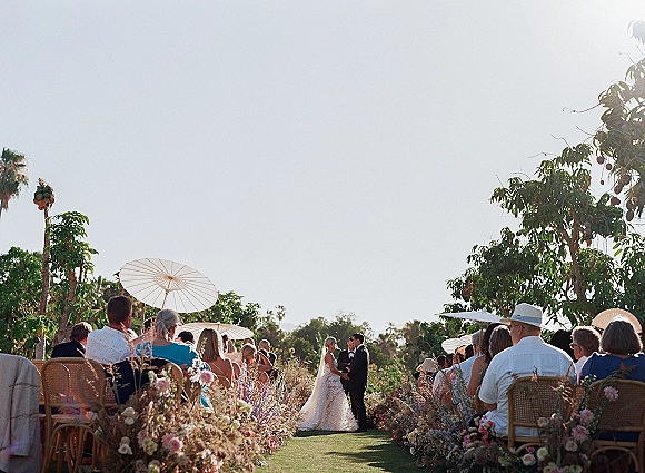 Wedding ceremony with bride in lace dress and veil and groom in black tuxedo at the altar, floral-lined aisle, guests with parasols in a garden