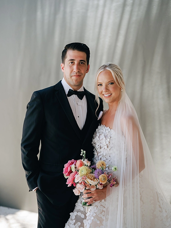 Couple portrait of bride in strapless gown with veil holding a colorful bouquet beside groom in black tuxedo against a soft neutral wall