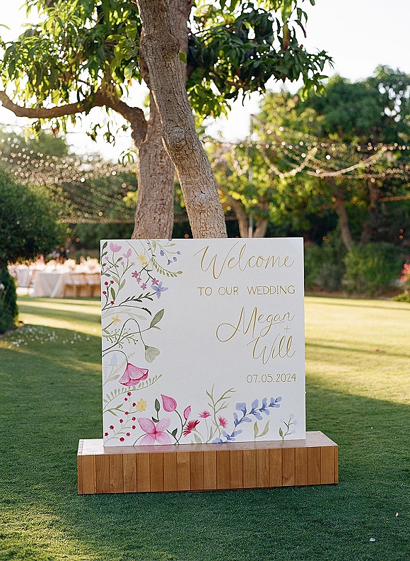 Wedding welcome sign with a painted floral illustration and gold calligraphy lettering on a white board, set on grass under string lights