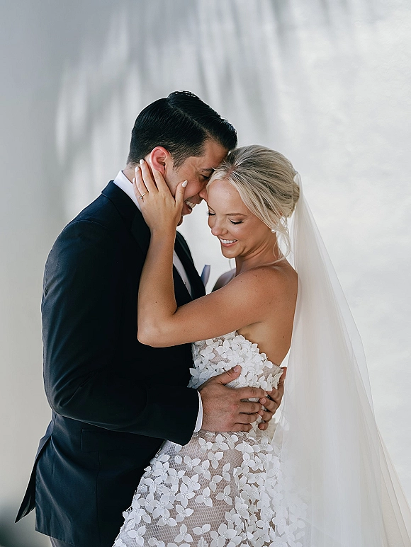 Couple portrait of bride and groom embrace, forehead touch in soft daylight, bride in veil and strapless lace dress by a white wall