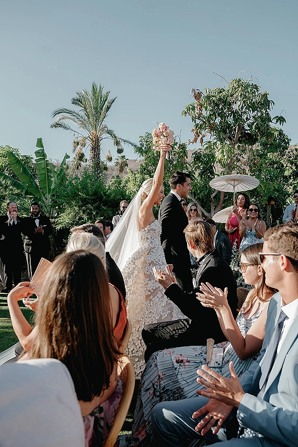 Wedding recessional as bride raising bouquet beside groom in tuxedo, long veil flowing while guests clap under white parasols in palm garden sky