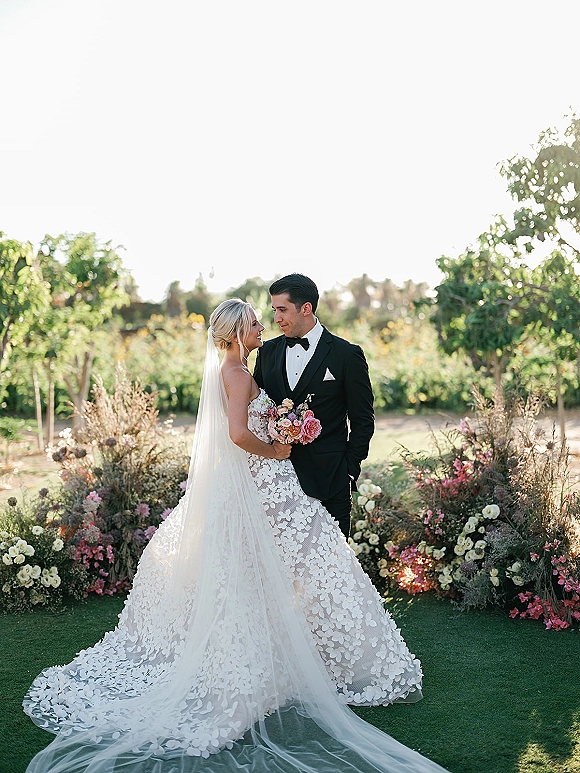 Couple portrait of bride and groom holding hands, she holds a bouquet with long veil trailing, in lush garden greenery and trees