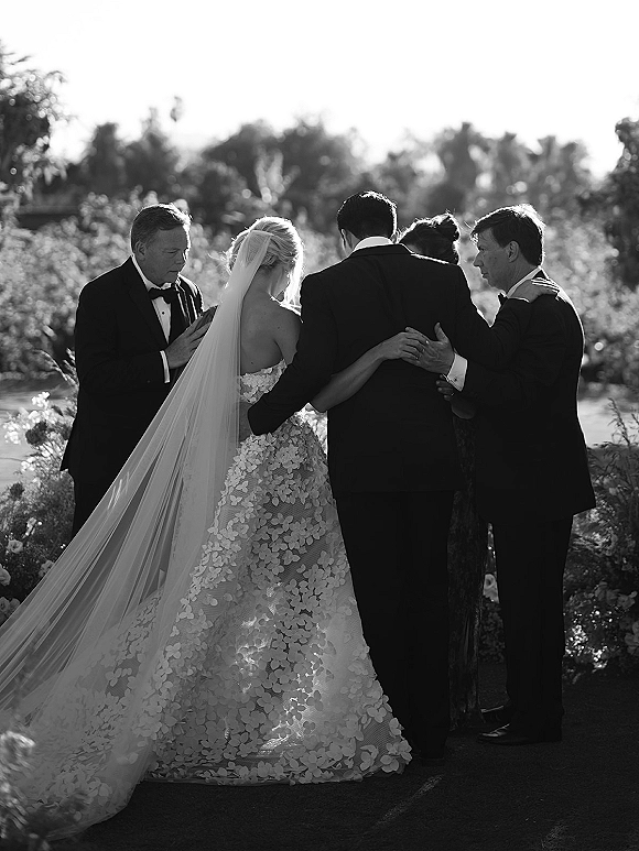 Ceremony moment with bride and groom holding hands as officiant leads an outdoor wedding ceremony in sunlit garden, veil flowing
