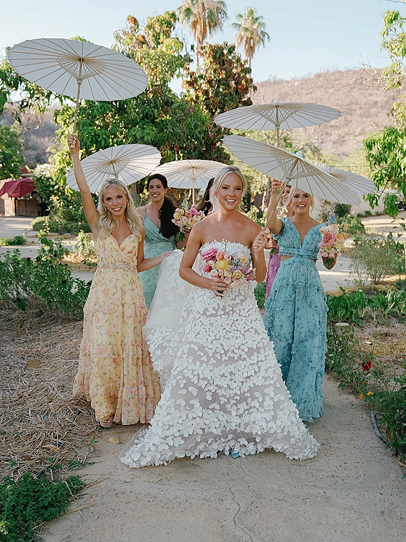 Bridesmaids portrait of bride with bridesmaids walking on a garden path, holding bouquets and white paper parasols under blue sky
