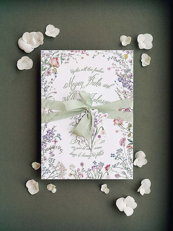 Wedding invitation with botanical wedding invitation floral illustrated border, tied with a sage green ribbon bow and scattered white petals on dark gray surface