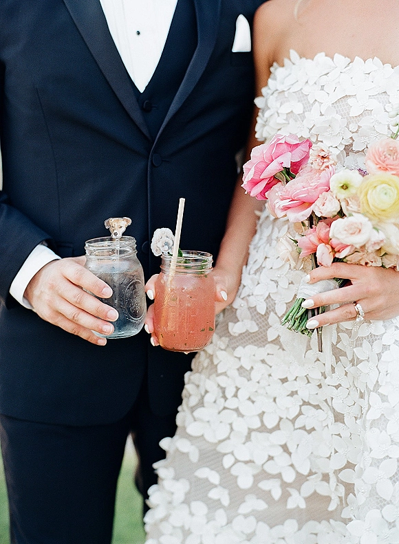 Newlywed portrait of bride and groom close up holding pink and peach bouquet and mason jar drinks amid softly blurred greenery background