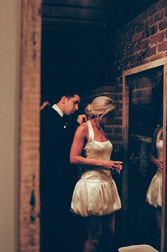 Couple portrait of bride and groom mirror moment as he adjusts her necklace, veil and pearl earrings by a brick wall indoors