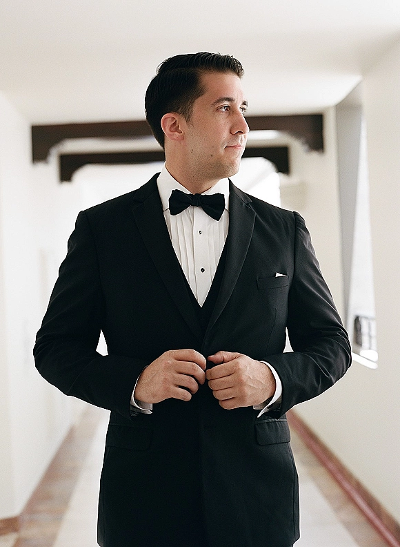 Groom portrait in a black tuxedo groom look, bow tie and pocket square, standing in a bright indoor hallway with windows and beams