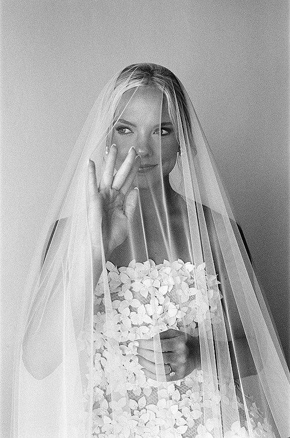 Bridal portrait, black and white bridal portrait of bride under veil in a strapless lace dress, showing wedding ring and manicured nails against a plain wall