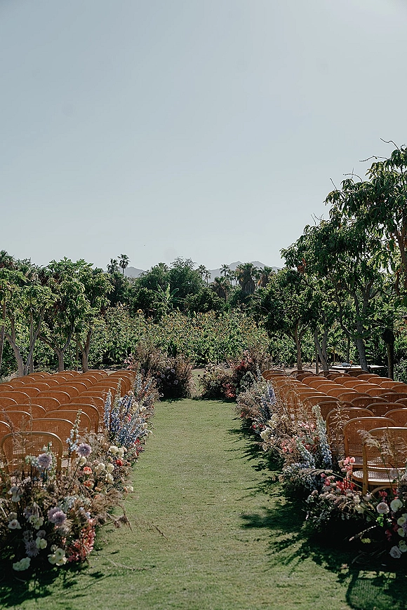Ceremony aisle decor with a grass runner and ground florals in clusters, flanked by wicker chairs under palms with mountains beyond