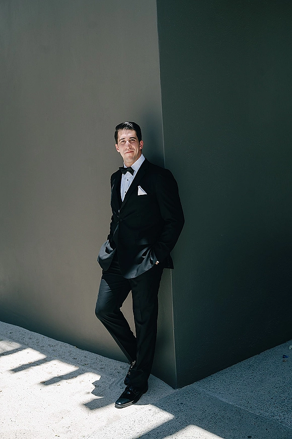 Groom portrait in a black tuxedo with bow tie and pocket square, standing on a concrete floor by a modern wall corner with shadows