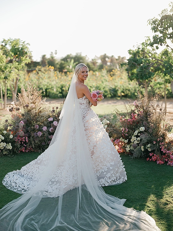 Bridal portrait of a bride in a strapless wedding dress with long veil, holding pink and peach rose bouquet in a garden setting