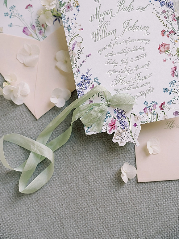 Wedding invitation suite with floral wedding invitations tied in sage green ribbon, vellum wrap and wax seal on gray linen with orchid petals