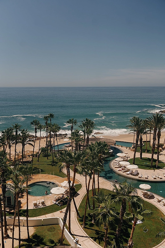 Beach resort view with oceanfront resort pool, curved swimming pool, palm trees, umbrellas and loungers overlooking waves and rocky coastline
